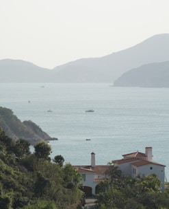 A peaceful coastal scene with a large villa in the foreground, surrounded by lush greenery. In the background, the calm sea stretches out towards a range of gently sloping hills. Several small boats are scattered across the water, contributing to the serene atmosphere.