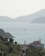 A peaceful coastal scene with a large villa in the foreground, surrounded by lush greenery. In the background, the calm sea stretches out towards a range of gently sloping hills. Several small boats are scattered across the water, contributing to the serene atmosphere.