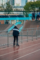 A person wearing inline skates and protective gear stands on a paved area near a railing, with a ramp in the background. Several other people are on top of the ramp, also equipped with skating gear. Trees and urban buildings are visible in the distance, creating an urban skate park environment.