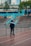 A person wearing inline skates and protective gear stands on a paved area near a railing, with a ramp in the background. Several other people are on top of the ramp, also equipped with skating gear. Trees and urban buildings are visible in the distance, creating an urban skate park environment.