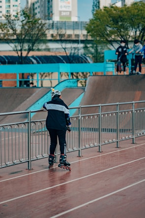 A person wearing inline skates and protective gear stands on a paved area near a railing, with a ramp in the background. Several other people are on top of the ramp, also equipped with skating gear. Trees and urban buildings are visible in the distance, creating an urban skate park environment.