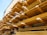 Multiple stacks of wooden planks are organized in horizontal rows on industrial metal shelving. The planks vary in size and are arranged systematically, creating a visually striking pattern with strong lines and angles. The background has a muted color, providing contrast to the wooden textures.