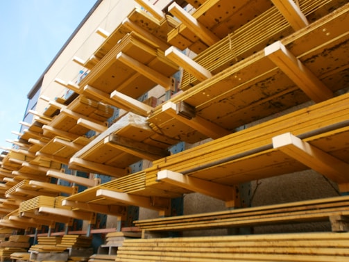 Multiple stacks of wooden planks are organized in horizontal rows on industrial metal shelving. The planks vary in size and are arranged systematically, creating a visually striking pattern with strong lines and angles. The background has a muted color, providing contrast to the wooden textures.
