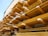 Multiple stacks of wooden planks are organized in horizontal rows on industrial metal shelving. The planks vary in size and are arranged systematically, creating a visually striking pattern with strong lines and angles. The background has a muted color, providing contrast to the wooden textures.