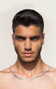 A young man with short dark hair and blue eyes is facing the camera against a plain, light-colored background. He has a short beard and is wearing a silver chain necklace around his neck.