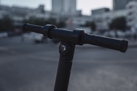 A close-up view of a black electric scooter handlebar in an urban environment. The background is blurred, showing indistinct buildings and a street scene, creating a bokeh effect.