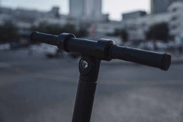 A close-up view of a black electric scooter handlebar in an urban environment. The background is blurred, showing indistinct buildings and a street scene, creating a bokeh effect.