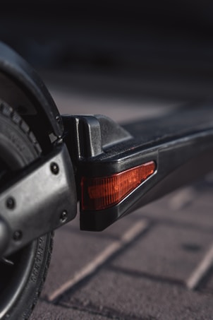 A close-up view of a scooter's rear section, highlighting the wheel and brake light. The tire is black with a sleek pattern, and the brake light features a translucent red cover. The scooter is positioned on a paved surface, and the lighting creates subtle shadows.