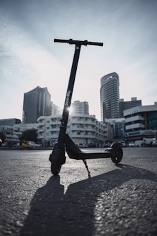 A black electric scooter is parked in an urban setting. The asphalt ground and modern high-rise buildings in the background create a metropolitan environment. The sky is overcast with a hint of sunlight peeking through.