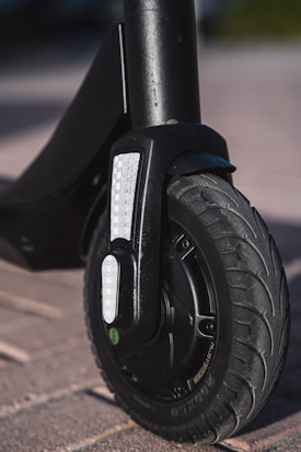 A close-up view of the front wheel of an electric scooter on a paved surface. The wheel features a tire with a detailed tread pattern, and there is a reflective strip attached to the fork.