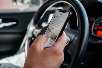 A person is holding a smartphone inside a car, with the phone displaying a tweet. The steering wheel and dashboard are visible, suggesting the person is a driver.