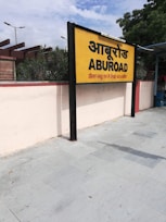 A yellow and black signboard prominently displays the name 'ABUROAD' in both Hindi and English, along with additional text in red below. The sign is positioned on a platform with a tiled floor. Greenery is visible in the background along with a blue sky partially covered by clouds.