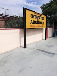 A yellow and black signboard prominently displays the name 'ABUROAD' in both Hindi and English, along with additional text in red below. The sign is positioned on a platform with a tiled floor. Greenery is visible in the background along with a blue sky partially covered by clouds.