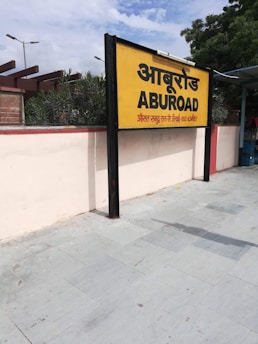 A yellow and black signboard prominently displays the name 'ABUROAD' in both Hindi and English, along with additional text in red below. The sign is positioned on a platform with a tiled floor. Greenery is visible in the background along with a blue sky partially covered by clouds.