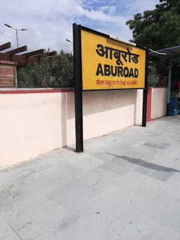 A yellow and black signboard prominently displays the name 'ABUROAD' in both Hindi and English, along with additional text in red below. The sign is positioned on a platform with a tiled floor. Greenery is visible in the background along with a blue sky partially covered by clouds.