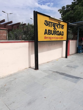 A yellow and black signboard prominently displays the name 'ABUROAD' in both Hindi and English, along with additional text in red below. The sign is positioned on a platform with a tiled floor. Greenery is visible in the background along with a blue sky partially covered by clouds.