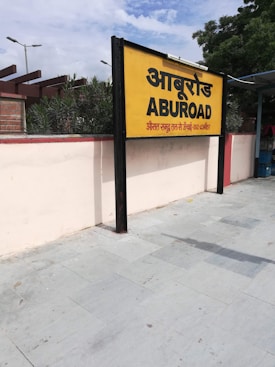 A yellow and black signboard prominently displays the name 'ABUROAD' in both Hindi and English, along with additional text in red below. The sign is positioned on a platform with a tiled floor. Greenery is visible in the background along with a blue sky partially covered by clouds.