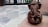 A small clay statue of the Hindu deity Ganesha is placed on a wooden surface. The statue features intricate details with a prominent elephant trunk, large ears, and a headdress. The background consists of a stone-tiled floor leading to a metal gate.