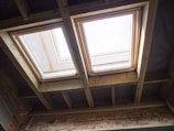 Two rectangular skylights are installed in a ceiling made of exposed wooden beams. Natural light streams through the windows, illuminating the wooden framework and a brick wall below. The sky is visible through the glass, suggesting a clear day.