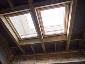 Two rectangular skylights are installed in a ceiling made of exposed wooden beams. Natural light streams through the windows, illuminating the wooden framework and a brick wall below. The sky is visible through the glass, suggesting a clear day.