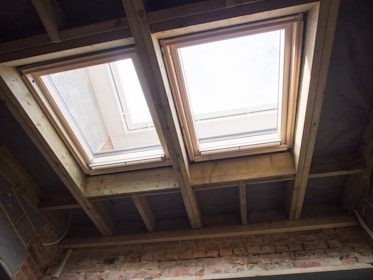 Two rectangular skylights are installed in a ceiling made of exposed wooden beams. Natural light streams through the windows, illuminating the wooden framework and a brick wall below. The sky is visible through the glass, suggesting a clear day.