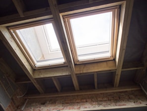Two rectangular skylights are installed in a ceiling made of exposed wooden beams. Natural light streams through the windows, illuminating the wooden framework and a brick wall below. The sky is visible through the glass, suggesting a clear day.