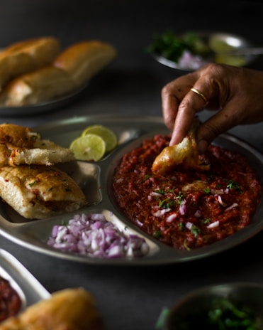 A hand is dipping a piece of bread into a rich, red curry garnished with fresh cilantro and diced onions on a metal plate. The plate also contains bread buns, lemon slices, and chopped onions. Additional plates with similar foods and garnishes can be seen in the background, hinting at a meal setting.