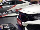 A close-up view of two sporty cars showcasing bold, aggressive designs with prominent branding. The focus is on the front side of a white car featuring a sleek grille and a red Honda badge, indicating its sporty model. In the background, another car exhibits dynamic graphics and branding.