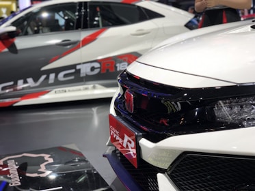 A close-up view of two sporty cars showcasing bold, aggressive designs with prominent branding. The focus is on the front side of a white car featuring a sleek grille and a red Honda badge, indicating its sporty model. In the background, another car exhibits dynamic graphics and branding.