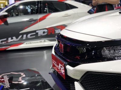 A close-up view of two sporty cars showcasing bold, aggressive designs with prominent branding. The focus is on the front side of a white car featuring a sleek grille and a red Honda badge, indicating its sporty model. In the background, another car exhibits dynamic graphics and branding.