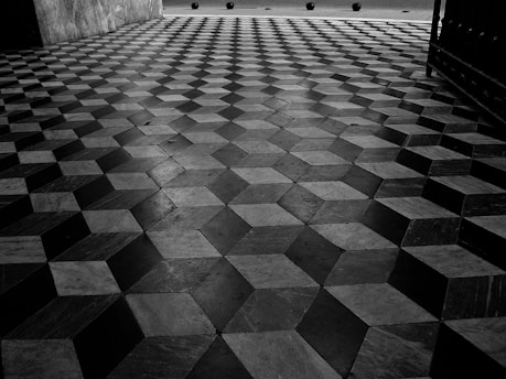 The image depicts a detailed pattern on a tiled floor featuring a geometric design with an optical illusion effect. The tiles are arranged in a way that creates the appearance of three-dimensional cubes. The background shows a few spherical objects, possibly bollards, placed at intervals.