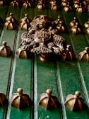 An ornate metal door with intricate detailing featuring a prominent central crest with a knight's helmet, flanked by decorative elements and surrounded by numerous rounded knobs with a gold finish.