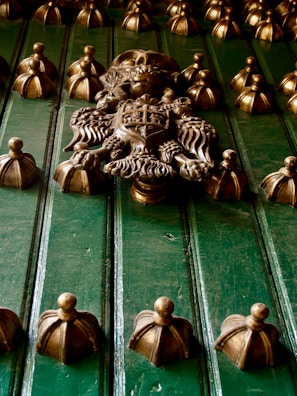 An ornate metal door with intricate detailing featuring a prominent central crest with a knight's helmet, flanked by decorative elements and surrounded by numerous rounded knobs with a gold finish.