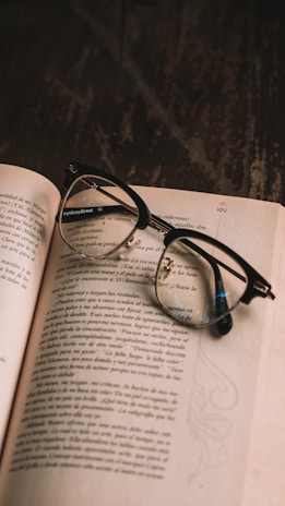 An open book with printed text in a foreign language lies on a dark wooden table. A pair of black eyeglasses with round lenses is placed on the book, partially covering the text.