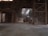 An industrial warehouse with a dirt floor, illuminated by natural light filtering through the roof. A construction vehicle, possibly a front loader, is parked inside, surrounded by large piles of sand or a similar material. The structure features metal beams and a high ceiling, creating a spacious yet enclosed atmosphere.