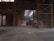An industrial warehouse with a dirt floor, illuminated by natural light filtering through the roof. A construction vehicle, possibly a front loader, is parked inside, surrounded by large piles of sand or a similar material. The structure features metal beams and a high ceiling, creating a spacious yet enclosed atmosphere.