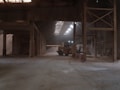 An industrial warehouse with a dirt floor, illuminated by natural light filtering through the roof. A construction vehicle, possibly a front loader, is parked inside, surrounded by large piles of sand or a similar material. The structure features metal beams and a high ceiling, creating a spacious yet enclosed atmosphere.