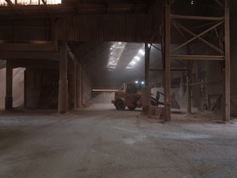 An industrial warehouse with a dirt floor, illuminated by natural light filtering through the roof. A construction vehicle, possibly a front loader, is parked inside, surrounded by large piles of sand or a similar material. The structure features metal beams and a high ceiling, creating a spacious yet enclosed atmosphere.