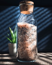 A glass jar with a cork lid filled with coarse pink salt and other spices is placed on a surface with shadow patterns from the blinds. In the background, there is a small potted plant with green leaves.