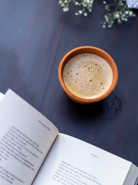 A cup of coffee rests on a dark wooden surface, next to an open book displaying poetry. Small white flowers are visible in the background, adding a delicate touch.