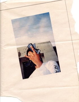 A person wearing a white long-sleeved shirt with printed text on the sleeve holds a stack of money in front of their face, obscuring their identity. They are standing against a background of a building with a partially clear blue sky.
