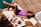 A person is sunbathing on a wooden deck beside a pool, lying on a yellow and white striped towel. They are wearing a purple swimsuit and sunglasses. Around them are a hat, a magazine, a bottle of sunblock, and a bowl of fruit with a drink.