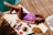 A person is sunbathing on a wooden deck beside a pool, lying on a yellow and white striped towel. They are wearing a purple swimsuit and sunglasses. Around them are a hat, a magazine, a bottle of sunblock, and a bowl of fruit with a drink.