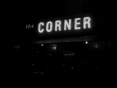 A neon sign with the word 'CORNER' illuminated in white light, with the surrounding area mostly dark. The full phrase on the sign appears to spell out 'the CORNER', as some letters are dimly visible around the brighter central word. Electric bulbs below the sign provide additional dim lighting to the scene.