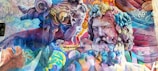 A vibrant mural featuring two classical-style sculptures interwoven with colorful graffiti art. The sculptures have intricate details with swirling hair and expressive faces, surrounded by layers of vivid graffiti text and patterns. The use of bright and bold colors enhances the dynamic contrast between the traditional sculptures and modern street art elements.
