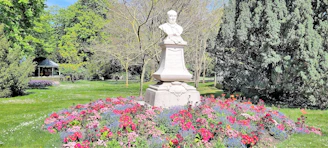 A stone statue of a person stands prominently in a garden surrounded by a vibrant array of flowers in various colors including red, pink, and purple. The garden features lush green grass and a variety of trees in the background, with a small gazebo visible on the left side. The scene is well-lit by daylight, creating patches of sunlight and shade.