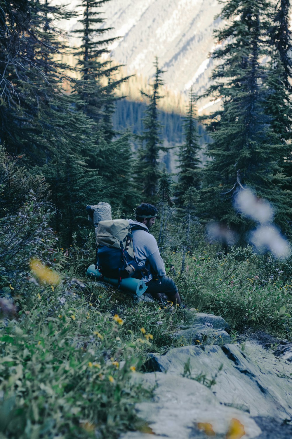 man in black jacket and blue backpack sitting on rock in front of green trees during