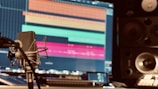 A professional audio recording setup with a microphone in the foreground, a large computer screen displaying a digital audio workstation, and two large studio speakers in the background. The screen shows various colored audio tracks, suggesting an ongoing music or sound editing project.