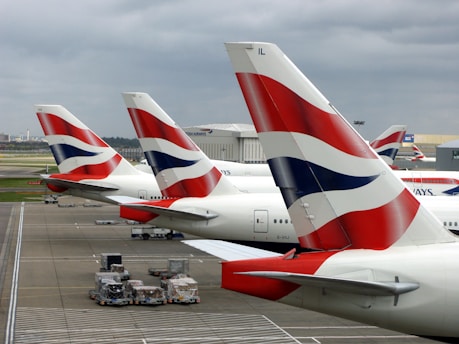 Several airplanes are parked at an airport terminal. The tails of the aircraft, adorned with a blue, white, and red design, are prominently visible. Pallets and airport ground handling equipment are positioned near the airplanes. The sky is overcast with gray clouds.