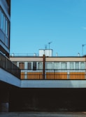 A section of urban architecture featuring a mix of modern and somewhat industrial design elements. The image captures a clear blue sky above a building with alternating orange and white window panels, covered by a balcony or walkway. Metal railings and an exposed brick section add to the linear aesthetic.